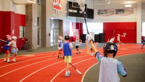 A young athlete in a helmet and jersey raises a lacrosse stick high on an indoor track, with other players practicing nearby.
