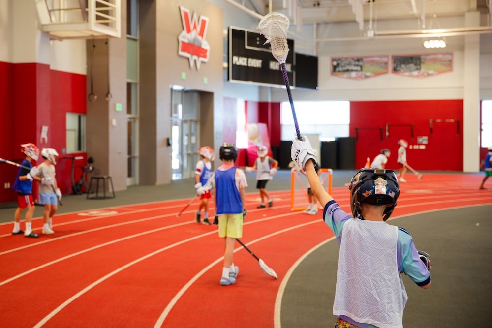 A young athlete in a helmet and jersey raises a lacrosse stick high on an indoor track, with other players practicing nearby.