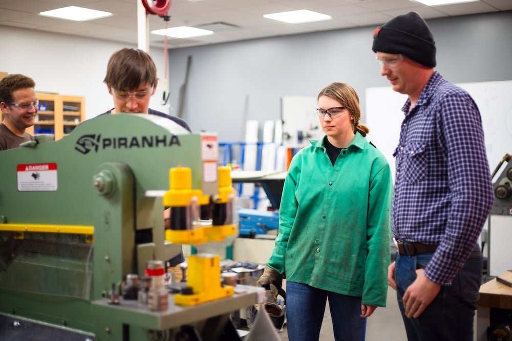 Four students stand around a machine in the Fabrication Shop and observe something.