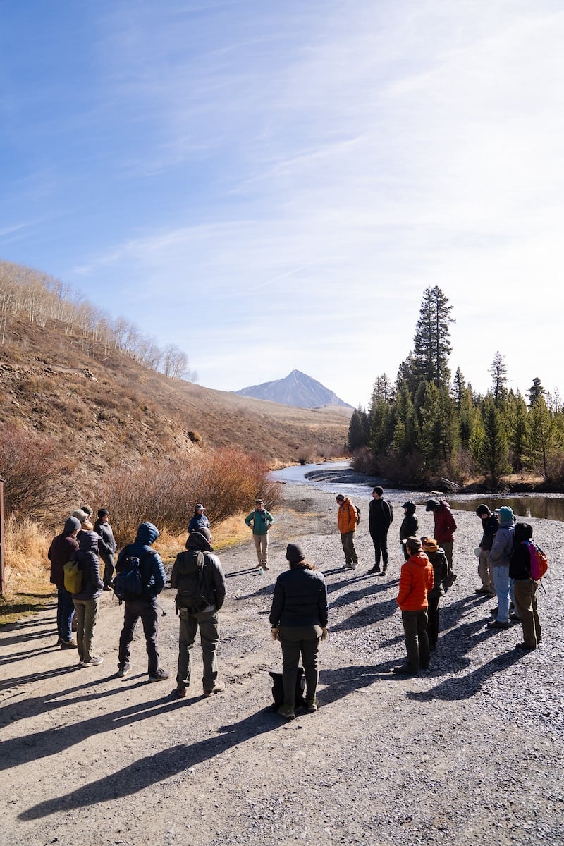 MEM faculty, students, and alumni listen to speakers from the Crested Butte Land Trust in a circle formation next to the Slate River in Crested Butte, Colorado, on a sunny day.