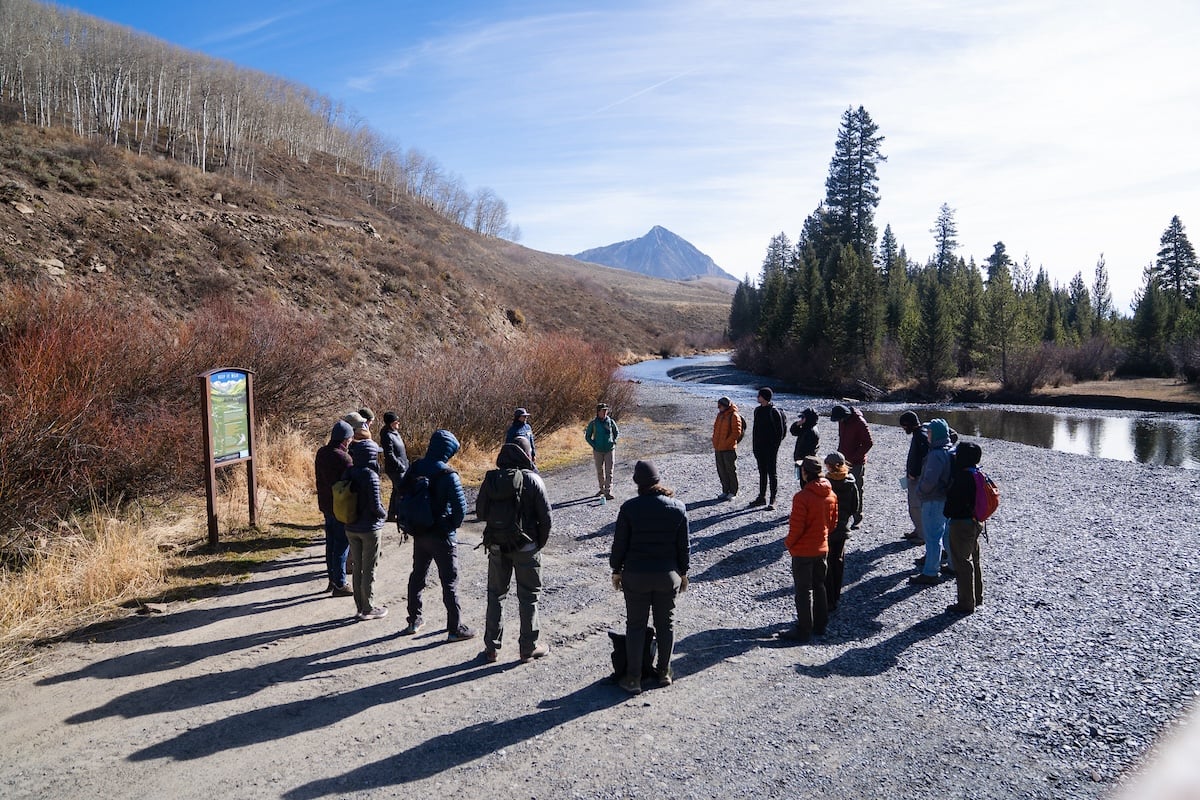 Mem faculty, students, and alumni listen to speakers from the Crested Butte Land Trust while standing in a circle formation next to the Slate River on a sunny fall day.