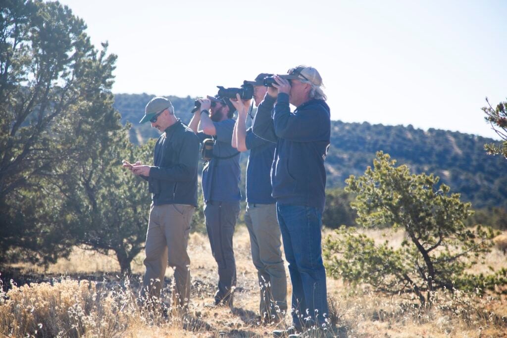 A group of students and Pat Magee stand and watch birds through binoculars and a camera lens during a field trip for an ornithology class.