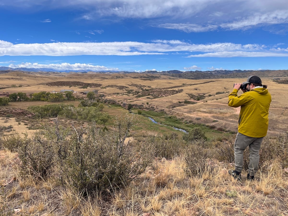 A man in a yellow jacket overlooks a valley with binoculars.