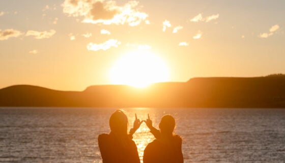 Two students put their fingers together to form a "w" while standing on the shore of Blue Mesa at sunset.