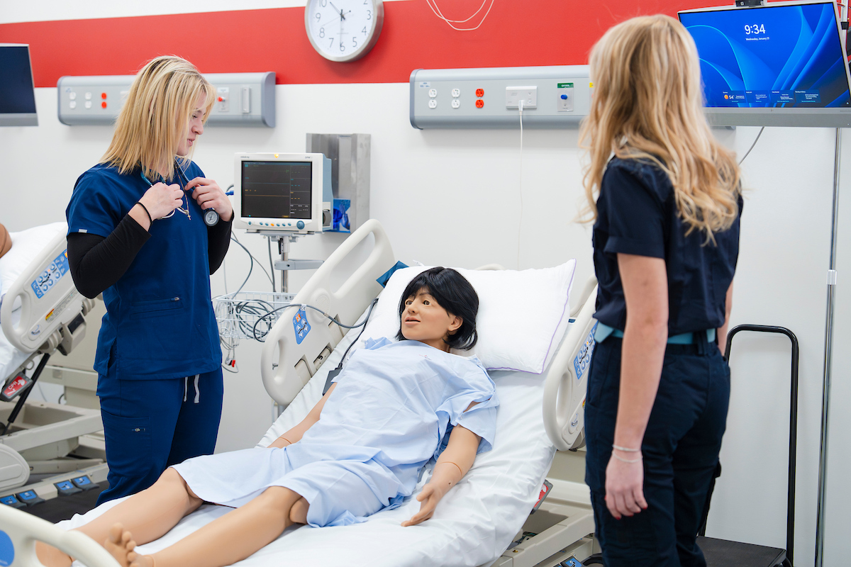 Two nursing students look down at a mannequin lying in a bed.