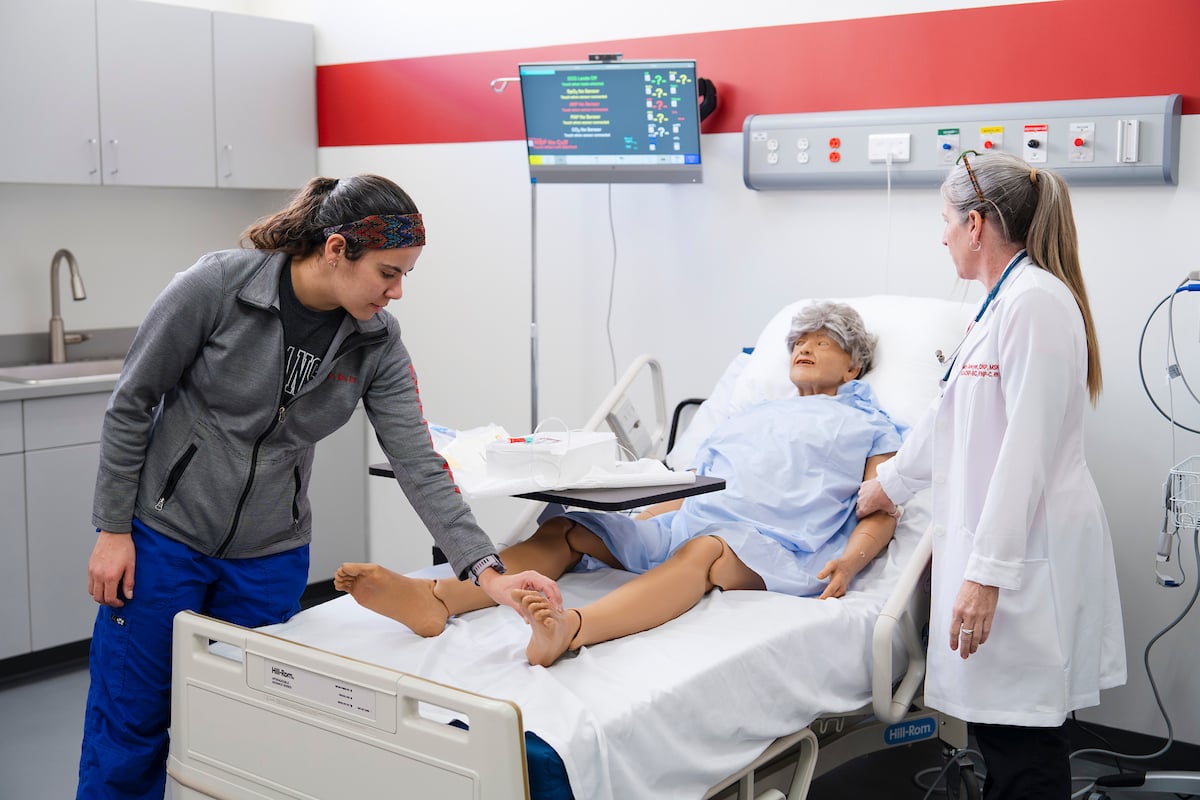 A nursing student feels for a pulse on a mannequin's foot in the simulation lab.