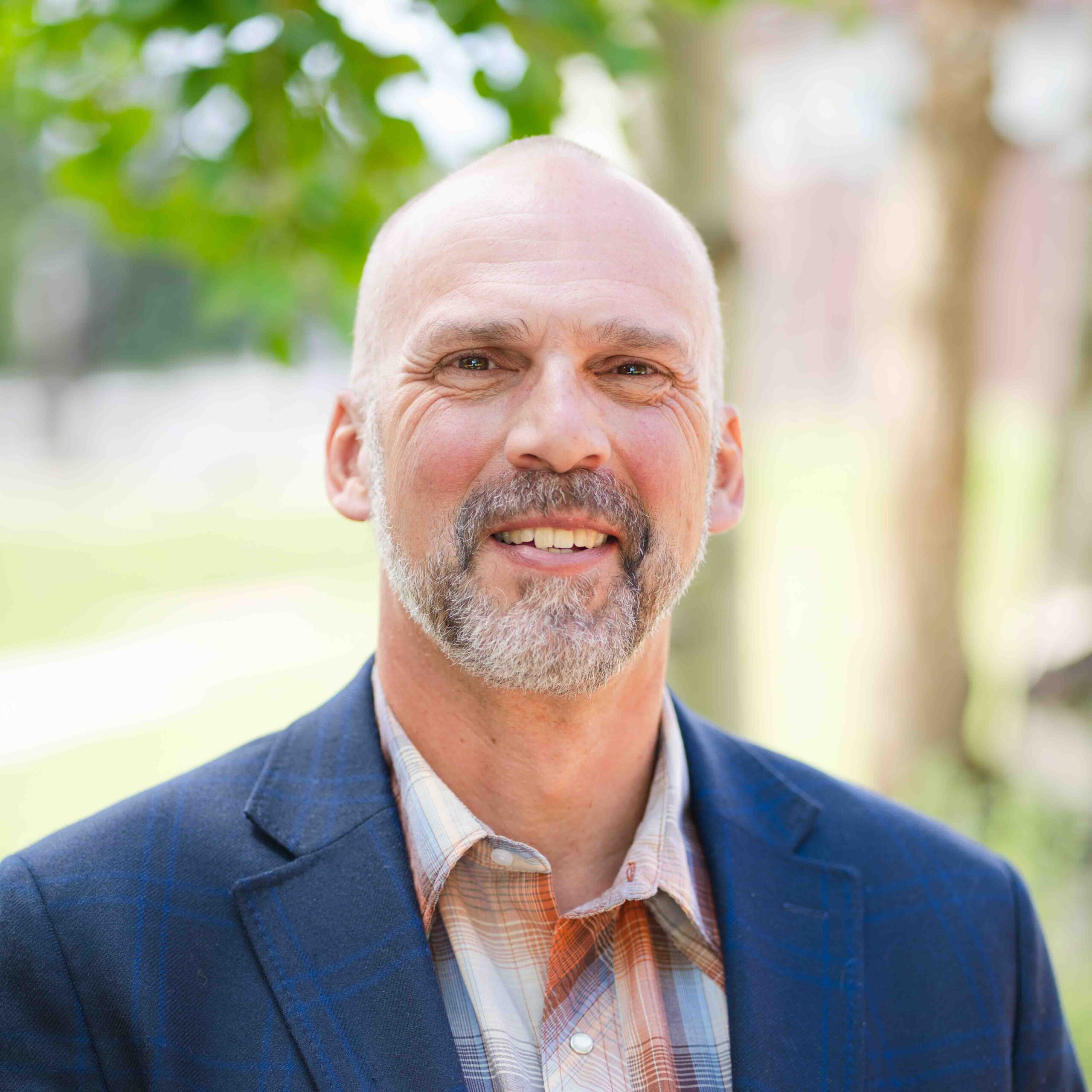 A bald individual with a salt-and-pepper beard smiles warmly while wearing a dark blue plaid blazer over a patterned button-up shirt, standing outdoors with green foliage in the background.