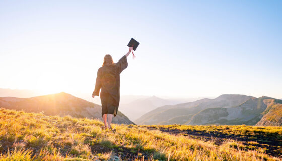 A student wearing a gown and raising her cap stands on a high alpine ridge at sunrise with big mountain views in the background.