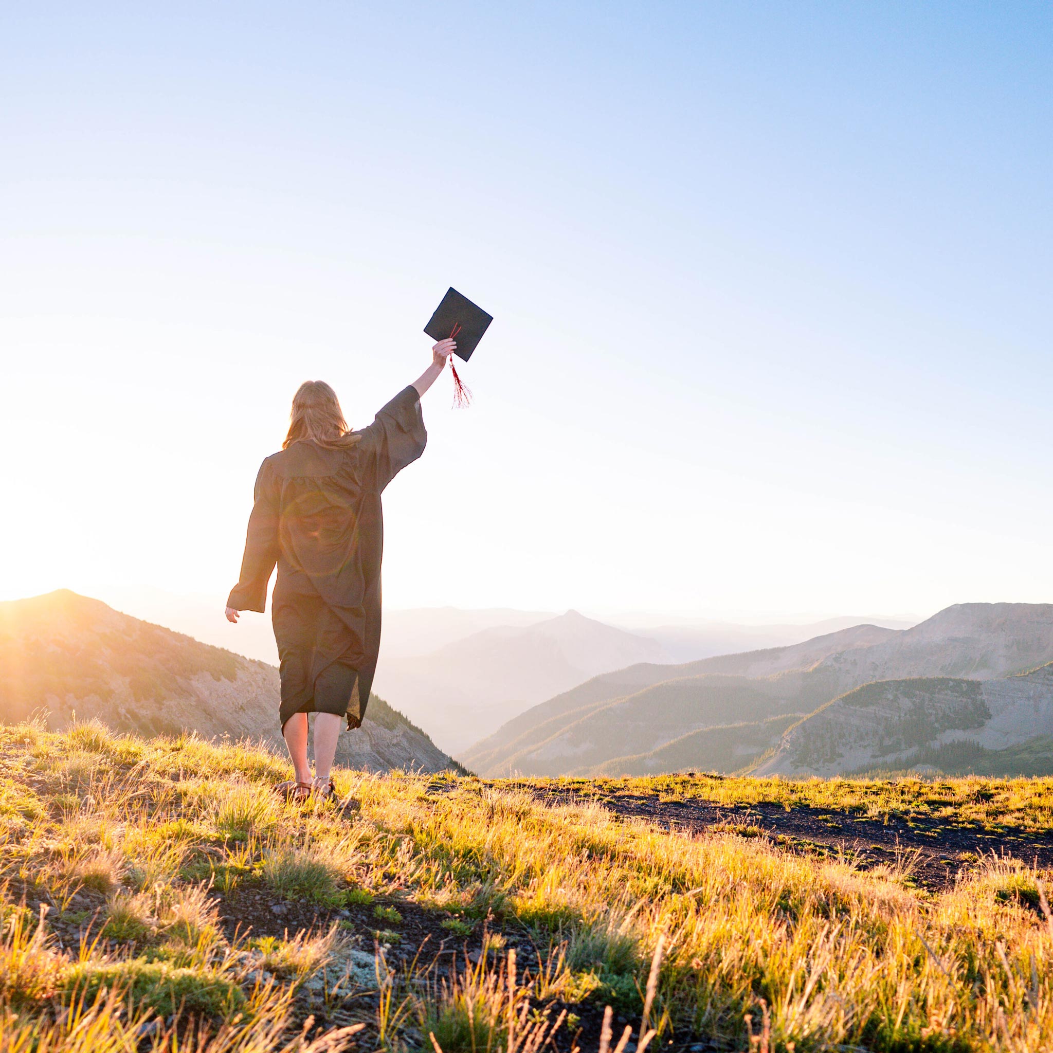 A student wearing a gown and raising her cap stands on a high alpine ridge at sunrise with big mountain views in the background.