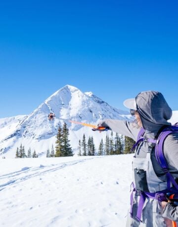 A girl wearing ski bibs and a backpack points at a triangular shaped mountain with her ski pole as she stands on a snowy alpine ridge on a blue sky day.