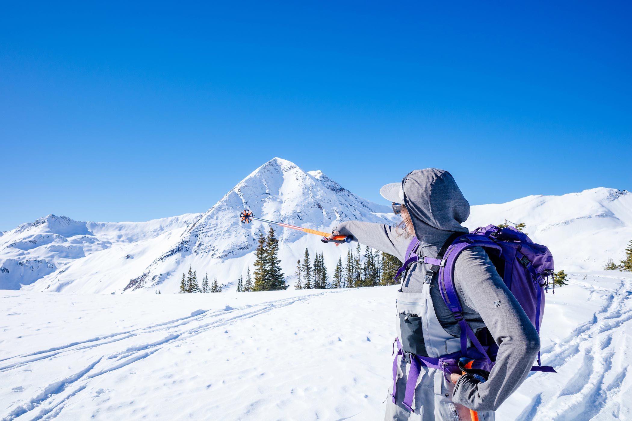 A girl wearing ski bibs and a backpack points at a triangular shaped mountain with her ski pole as she stands on a snowy alpine ridge on a blue sky day.