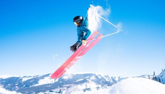 A skier grabs her skis while jumping in the air on a blue sky day with mountains in the background.