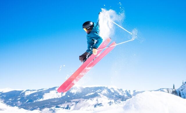 A skier grabs her skis while jumping in the air on a blue sky day with mountains in the background.