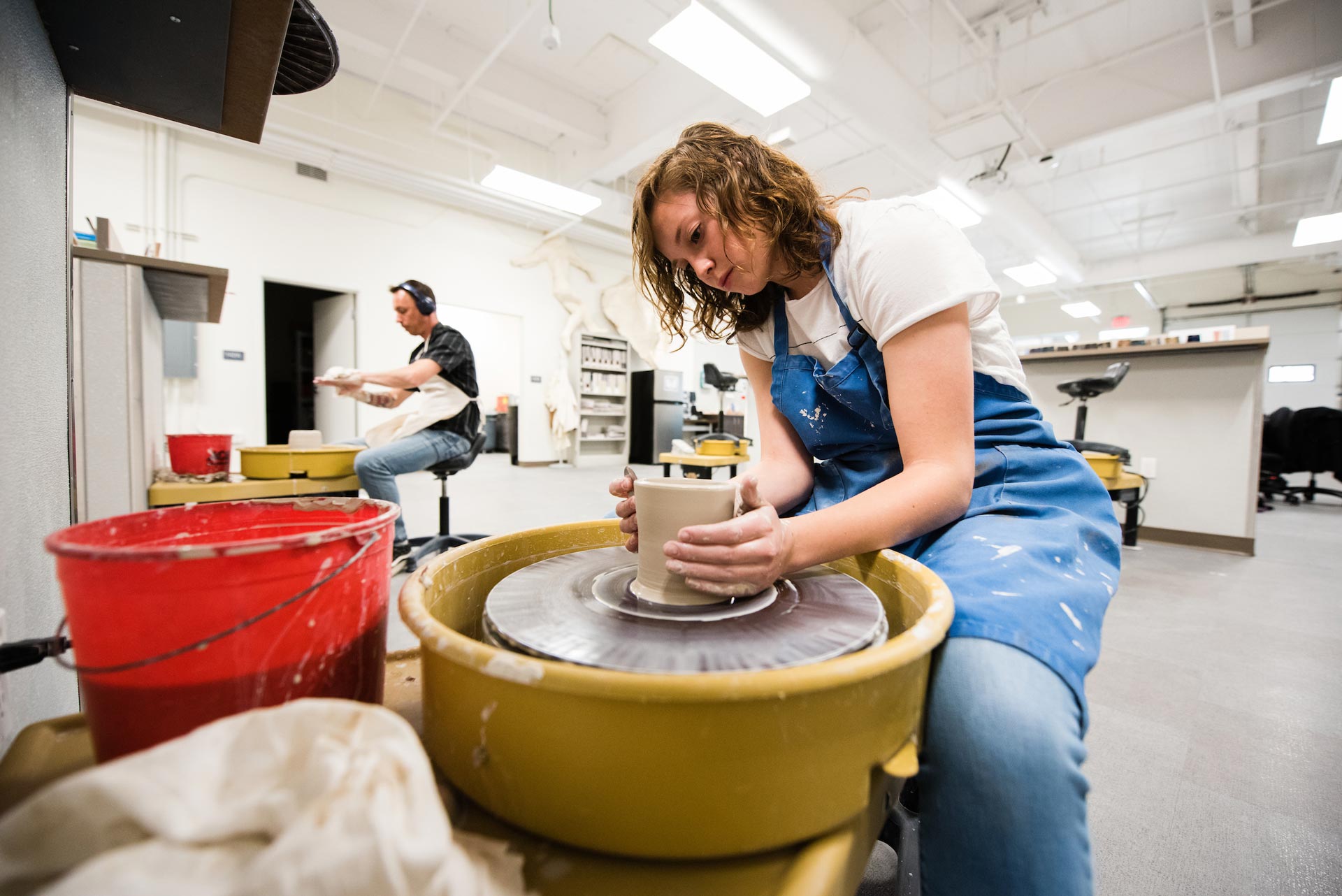 A student wearing an apron sits as she spins a pot on the pottery wheel.
