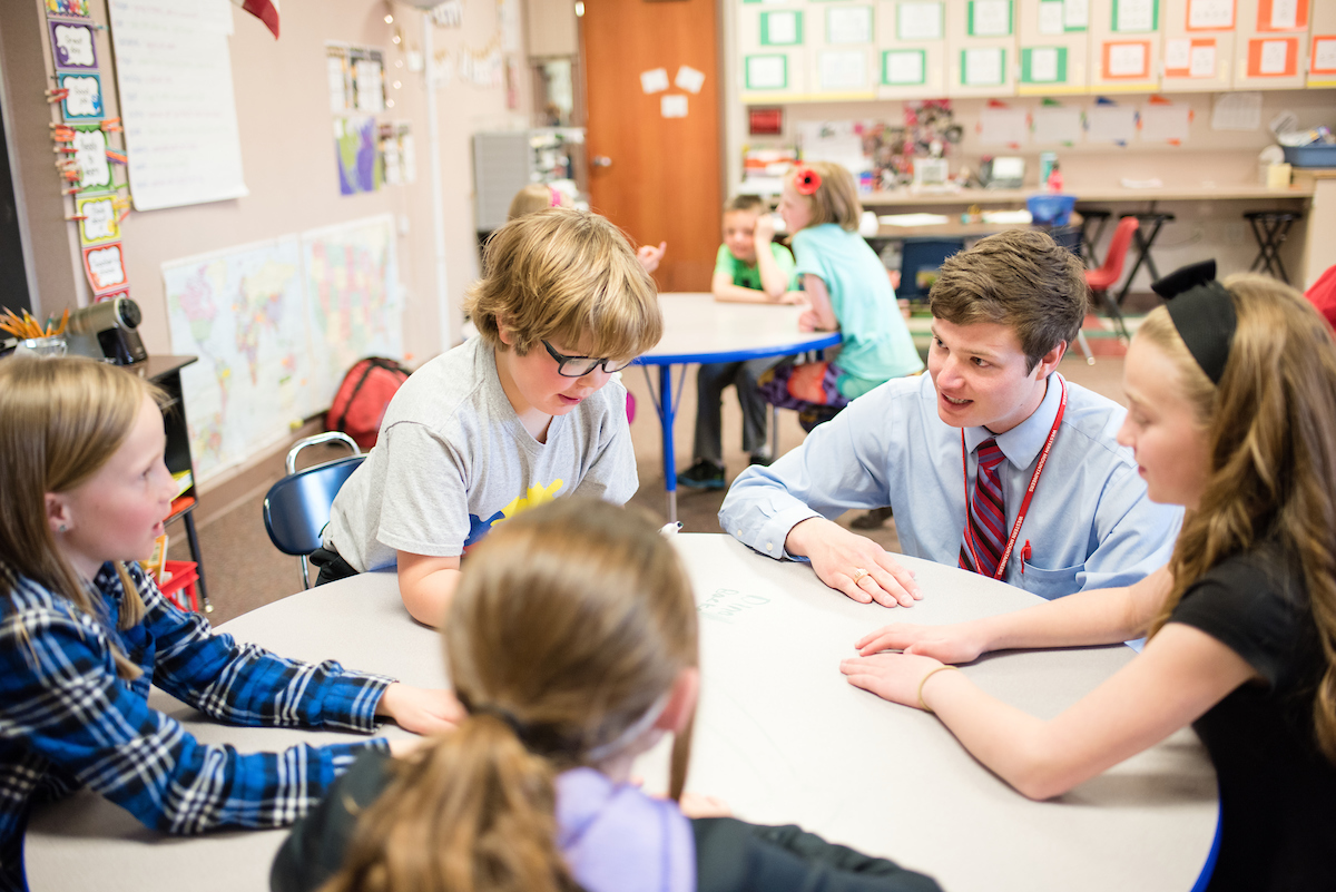 A male student teacher helps kids learn around a table.