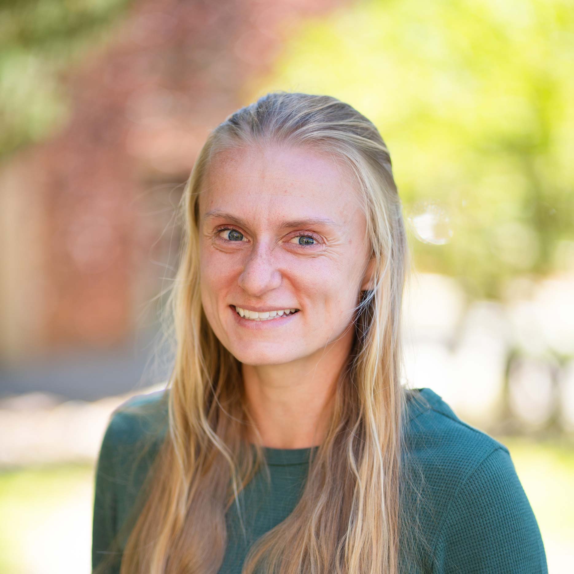 A person with long blonde hair smiles warmly while wearing a green long-sleeved top outdoors.