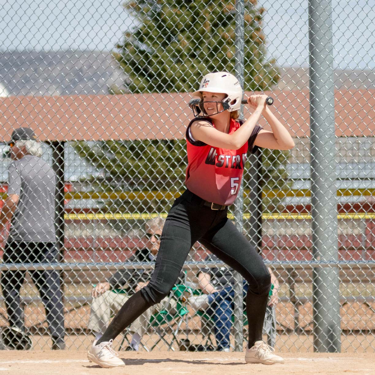 Linnea Baldner prepares to bat during a softball game. She faces the camera as she looks towards the pitcher. She has the bat raised over her left shoulder.