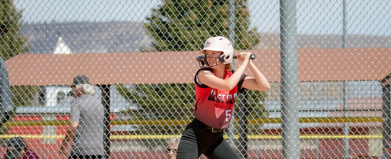 Linnea Baldner prepares to bat during a softball game. She faces the camera as she looks towards the pitcher. She has the bat raised over her left shoulder.