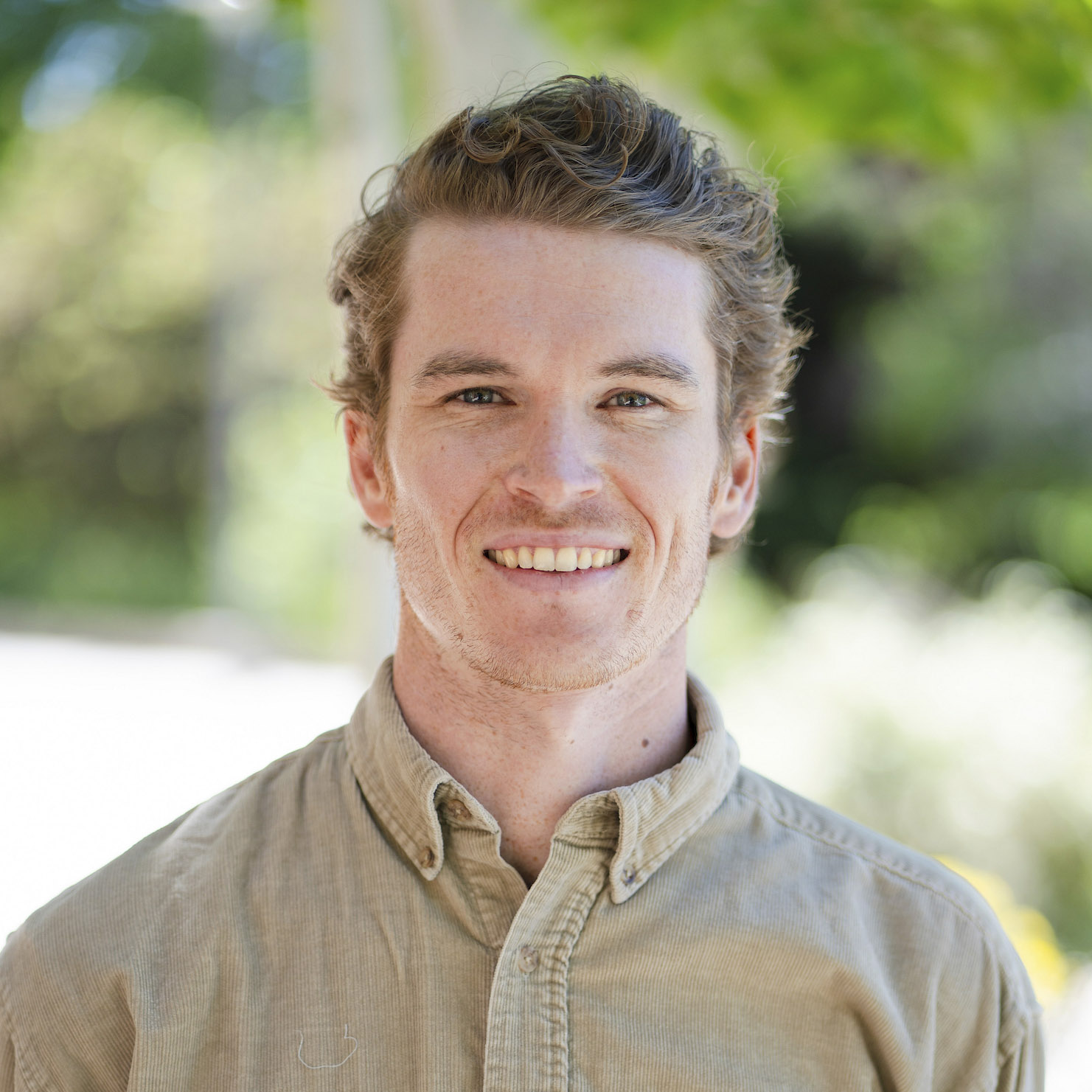 A smiling person wears a beige corduroy shirt outdoors against a blurred green background.
