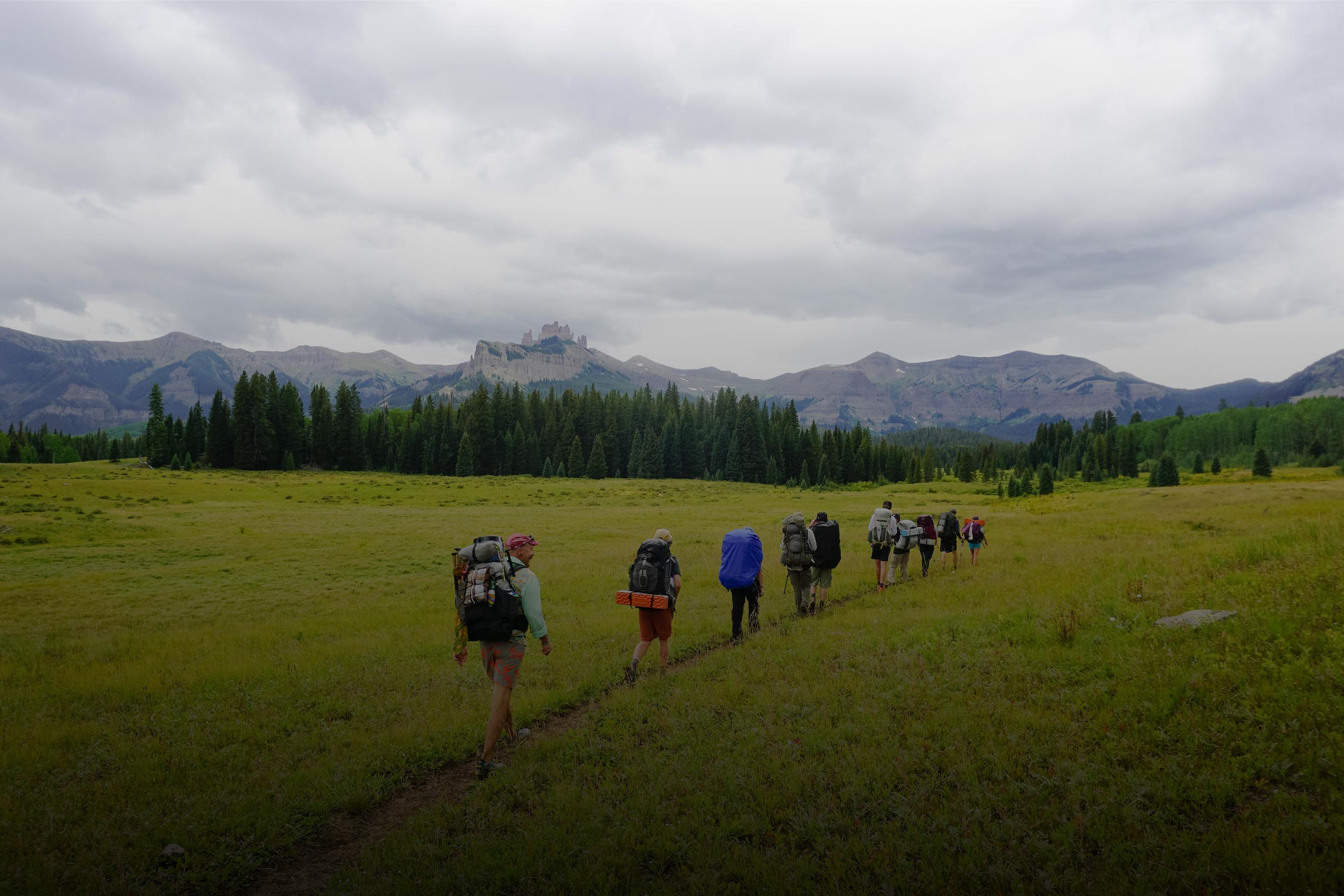 An ROE land-based class in single-file backpacks up a flat mountain meadow with the Castles and West Elk Peaks in the background.