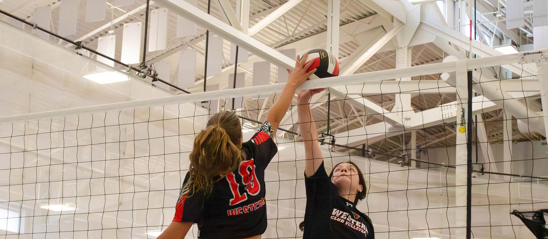 Two Western Women's Volleyball players jump up next to either side of the net to block the ball. They both have a hand on either side of the volleyball.