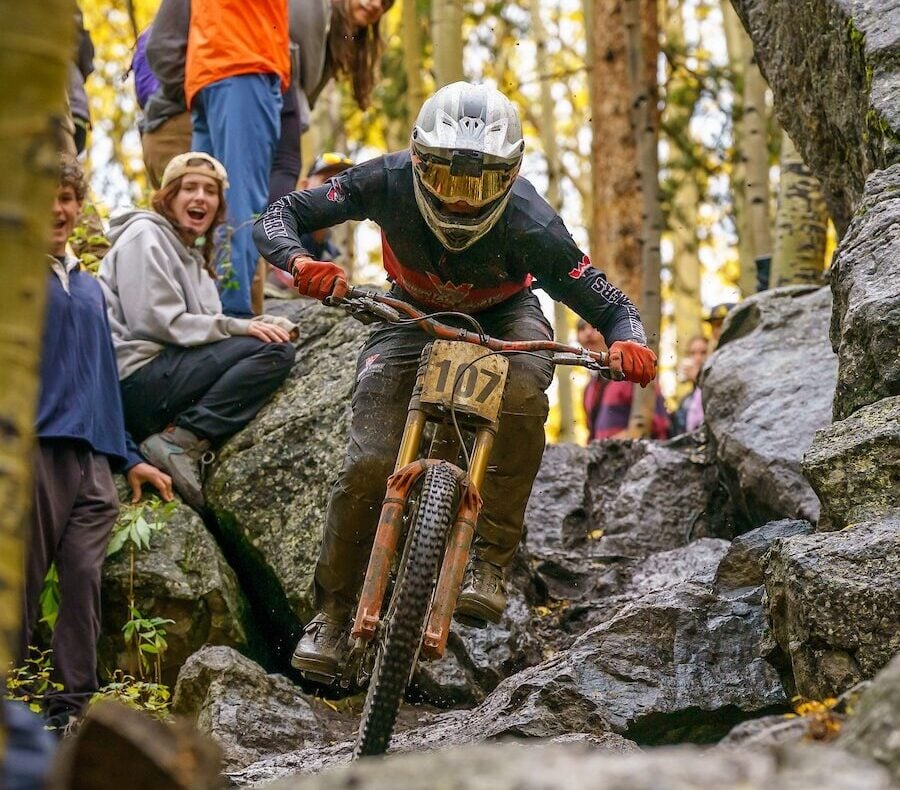 Mountain biker (in the part of the downhill corse that is in the trees) rides down large rocky terrain. Spectates stand on rocks around the course in awe
