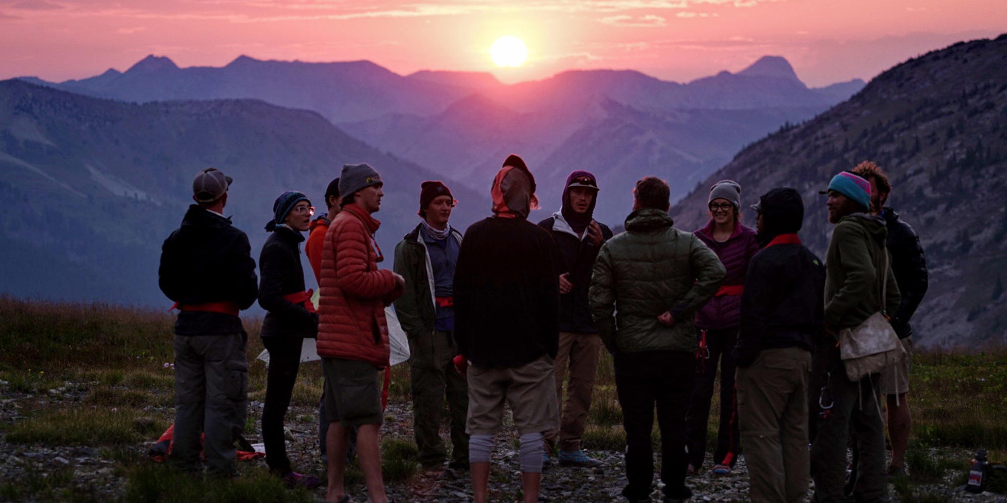 Group of students talking during a hike with the sun setting behind the mountains in the background
