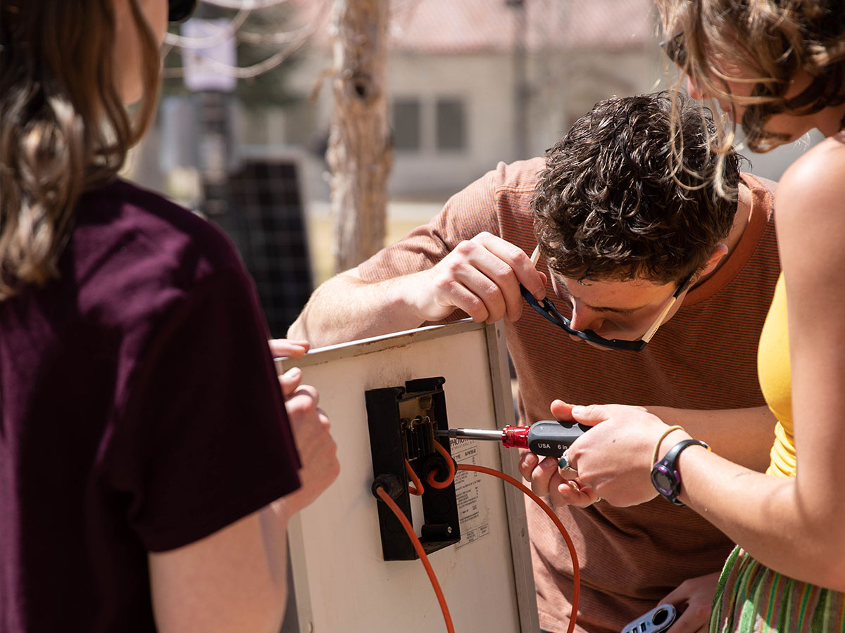 A student uses a screwdriver to remove a junction box on the back of a solar panel while two other students hold the panel up