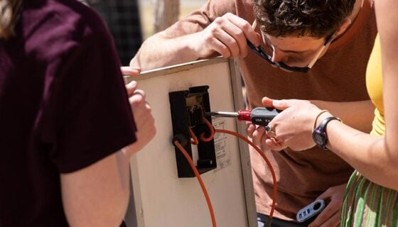 A student uses a screwdriver to remove a junction box on the back of a solar panel while two other students hold the panel up