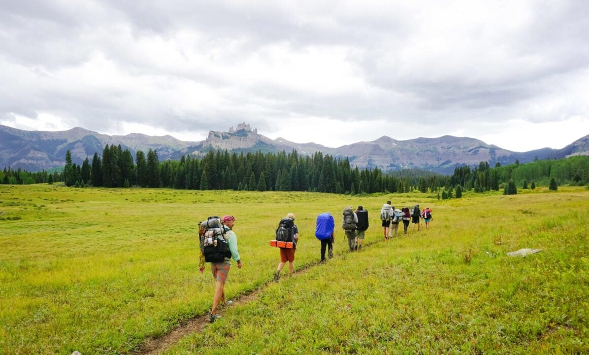 An ROE land-based class in single-file backpacks up a flat mountain meadow with the Castles and West Elk Peaks in the background.