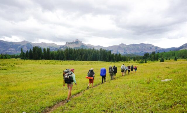 An ROE land-based class in single-file backpacks up a flat mountain meadow with the Castles and West Elk Peaks in the background.