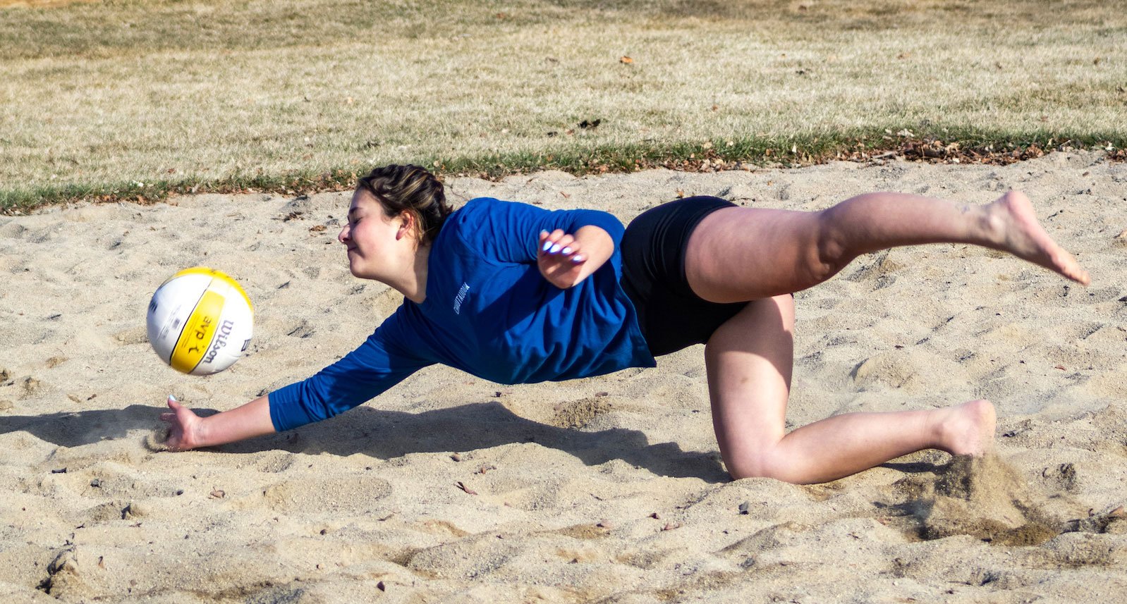 A student dives for a volleyball on a sand court.