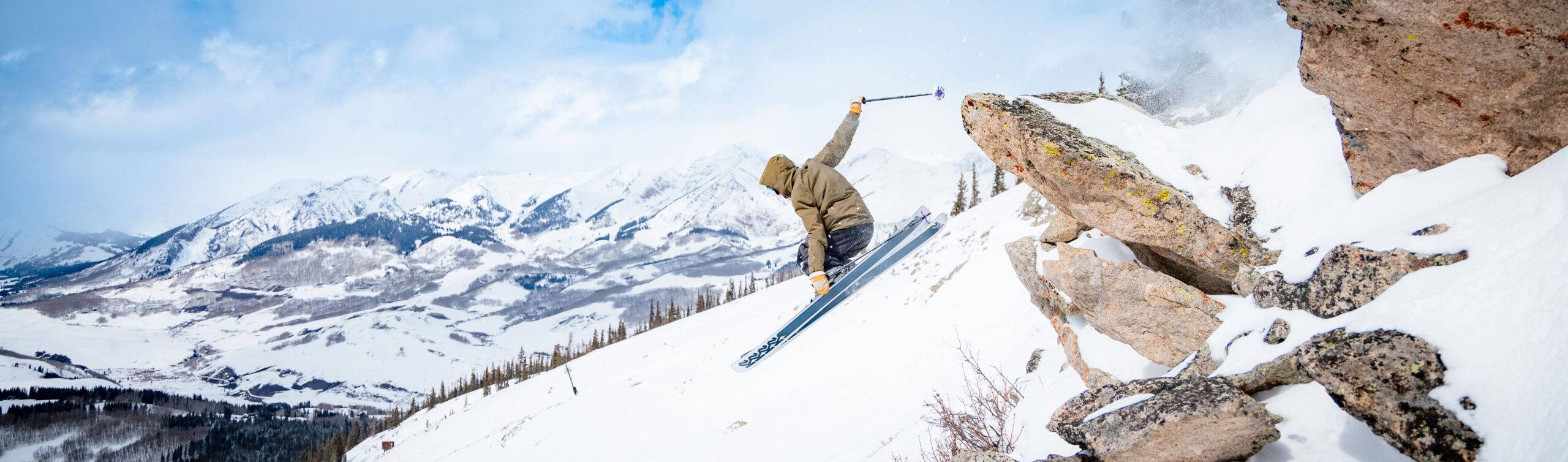 A skier airs off a cliff while grabbing his skis with mountain views in the background.