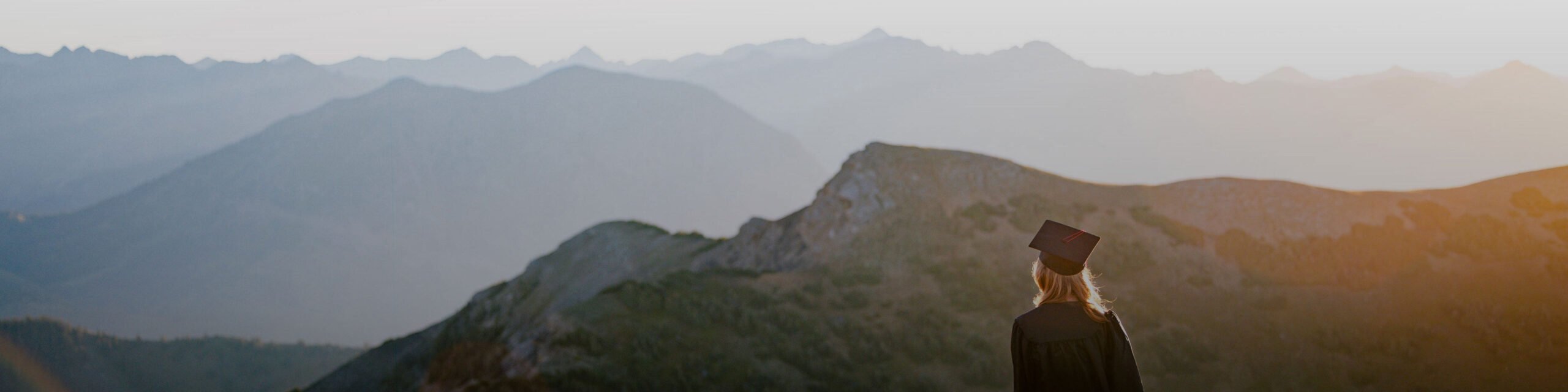 A student wearing a graduation cap and gown stands on a high alpine ridge at sunrise.