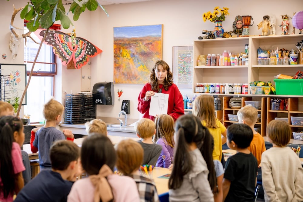 A student teacher holds up a coloring sheet in front of the first grade art class.