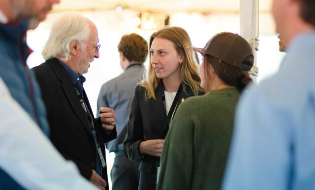 A group of people engage in conversation under a tent, with one person in a black blazer and another in a green sweater.