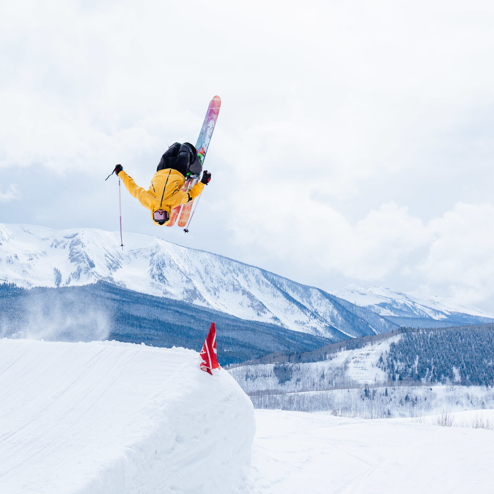 A skier does a backflip off a park jump with mountains in the background.