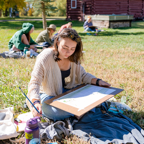 A student sits on grass and watercolor paints a scene with a barn and fall colors behind her.