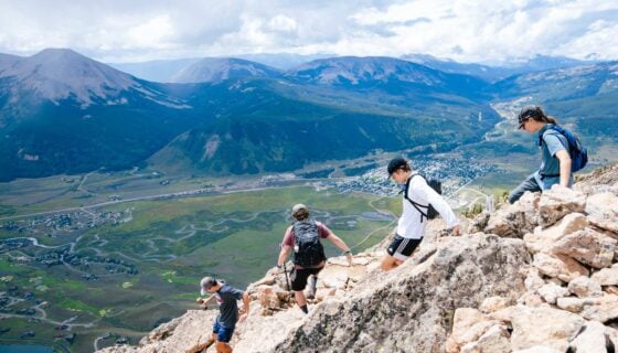 Four students wearing backpacks descend a rocky section of Mt. Crested Butte with big mountain views in the background.