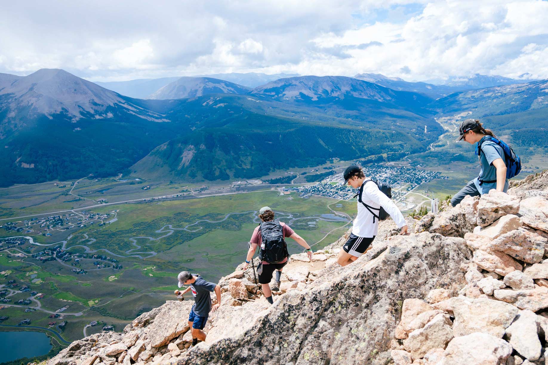 Four students wearing backpacks descend a rocky section of Mt. Crested Butte with big mountain views in the background.