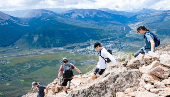 Four students wearing backpacks descend a rocky section of Mt. Crested Butte with big mountain views in the background.