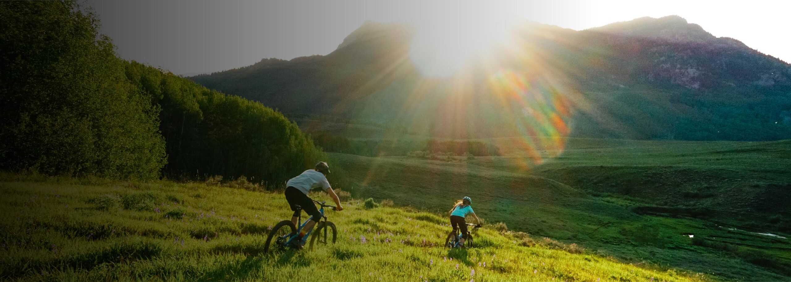 Two bikers descend a grassy hill at sunset.