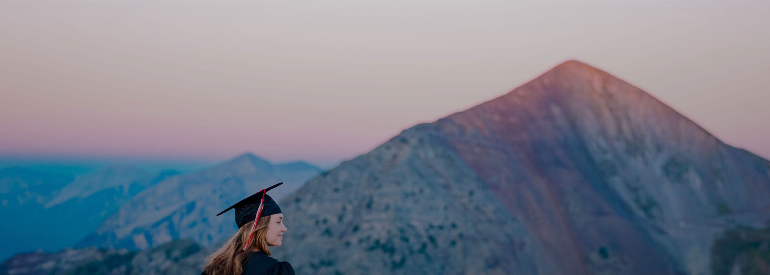 A student, looking off into the distance, wears a cap and gown as she stands on a high alpine ridge with a large peak behind her at sunrise.