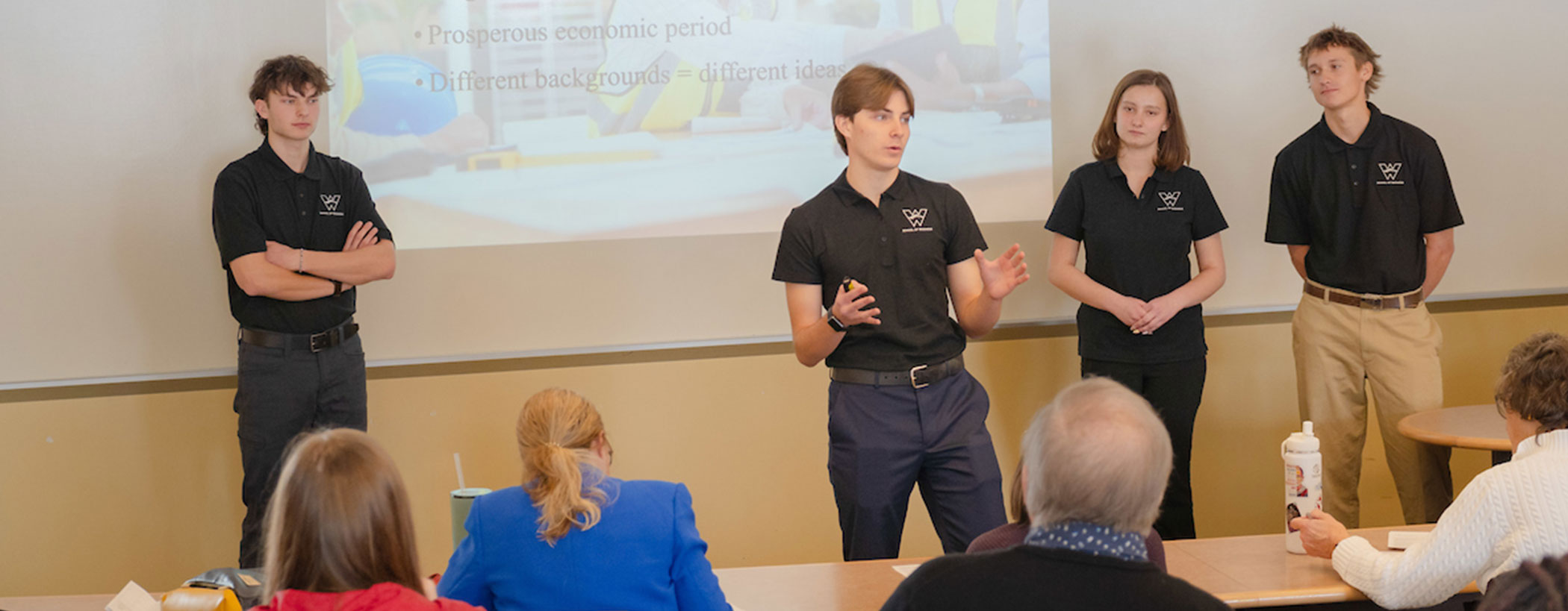 Borick Scholars present their proposal to the judges. Three young men and one young lady present as a team in the front of a classroom with projected slides behind them.