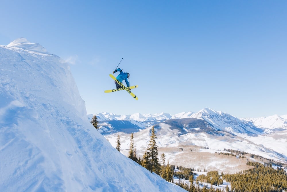 A skier jumps off the Crested Butte classic hit, Honey Pie while grabbing mute. Gothic mountain can be seen in the background with blue skies.